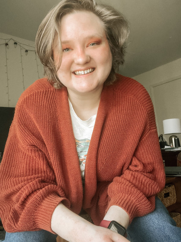 Elliot, a white nonbinary person, sits facing the camera. They are wearing blue jeans and a rust-red sweater.