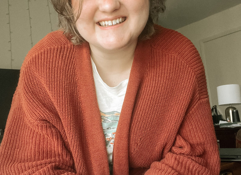 Elliot, a white nonbinary person, sits facing the camera. They are wearing blue jeans and a rust-red sweater.