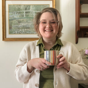 Elliot - a white nonbinary person with dirty blond hair and clear framed glasses - stands in front of a white wall with a Monet painting on it. They're wearing a green polo shirt and white cardigan, and they're holding a tan mug with vertical rainbow stripes.